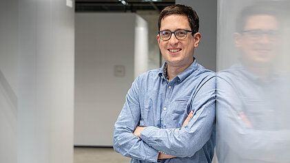  Ein Mann mit Brille lehnt an einer Wand und blickt lächelnd in die Kamera.