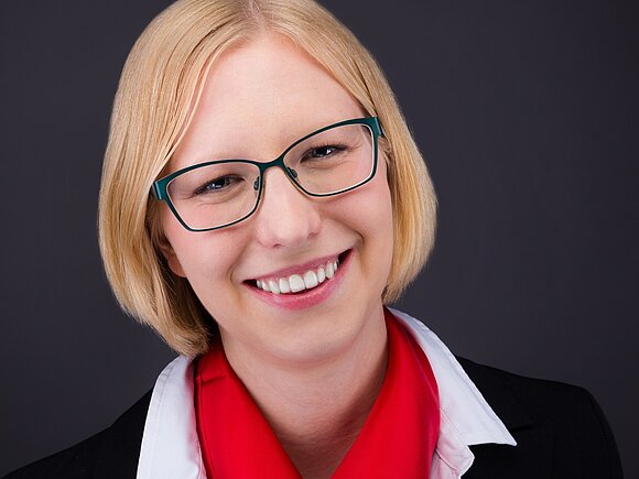 <br /> Eine Frau mit Brille und rotem Schal, die freundlich in die Kamera lächelt.